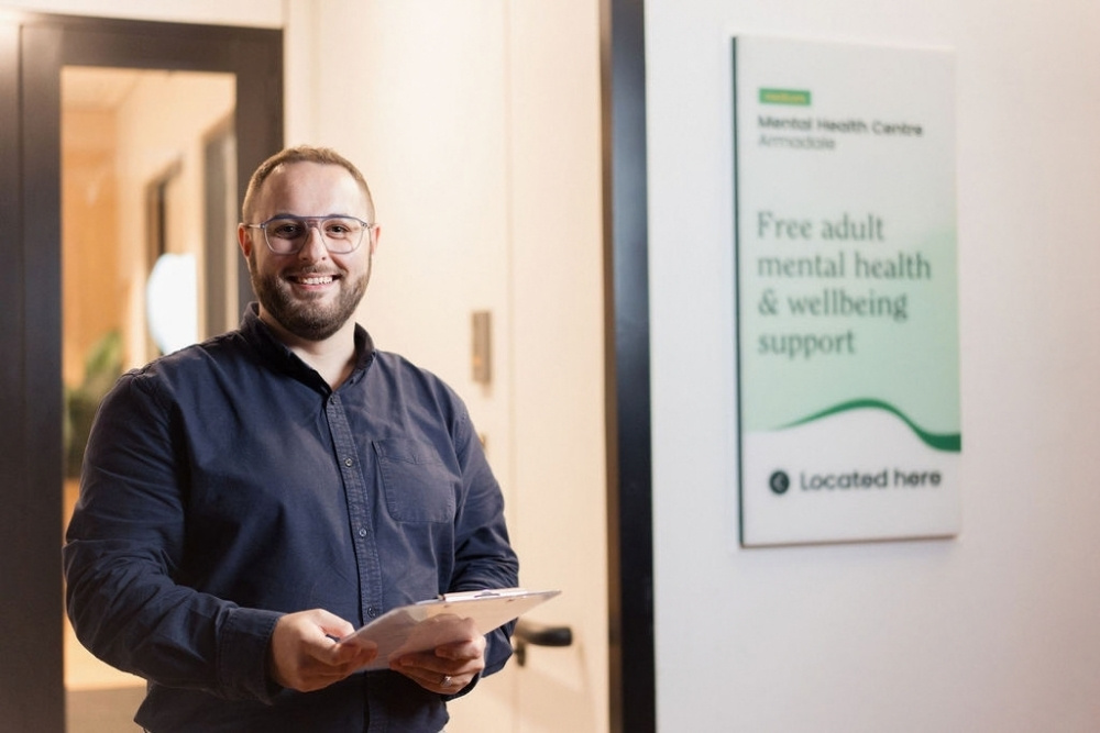 A smiling staff member stands with a clipboard outside a Medicare Mental Health Centre
