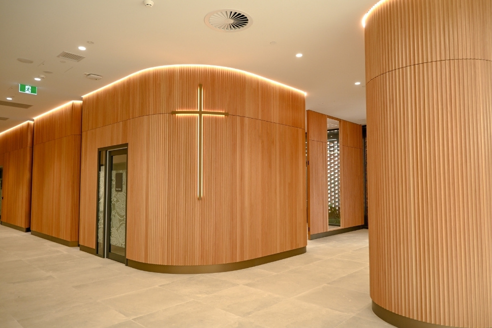 A large timber cross, illuminated with a warm golden glow, provides a welcoming focal point at the entrance of the St John of God Midland Private Hospital chapel