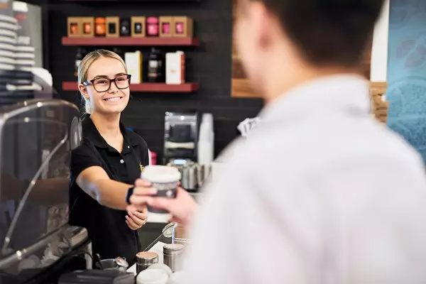 A female barista with glasses hands a coffee to a male customer from behind the counter